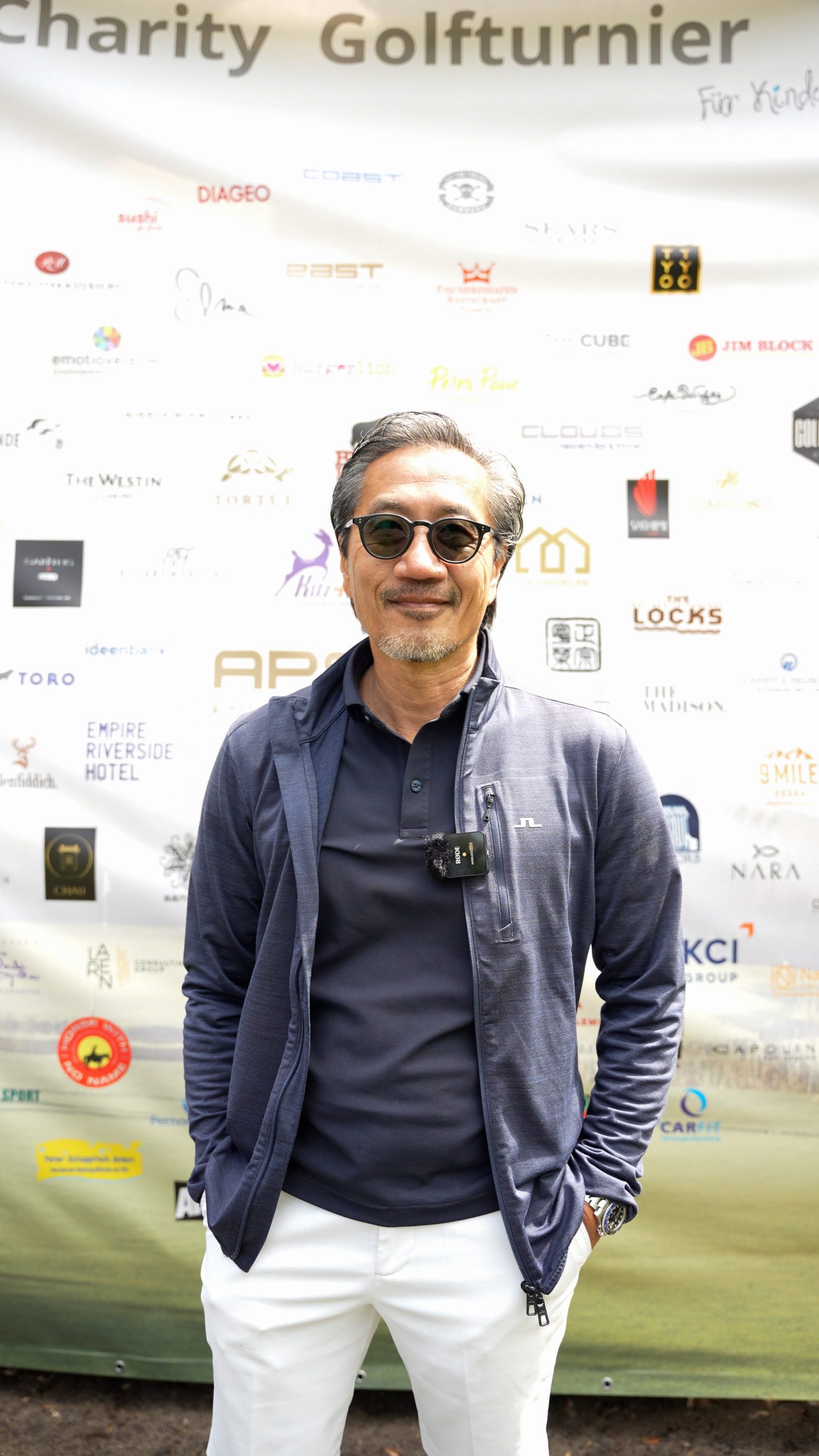 Der Geschäftsführer von APS Bar und Supply, Shang You steht vor der Sponsorenwand des Benefiz-Golfturniers