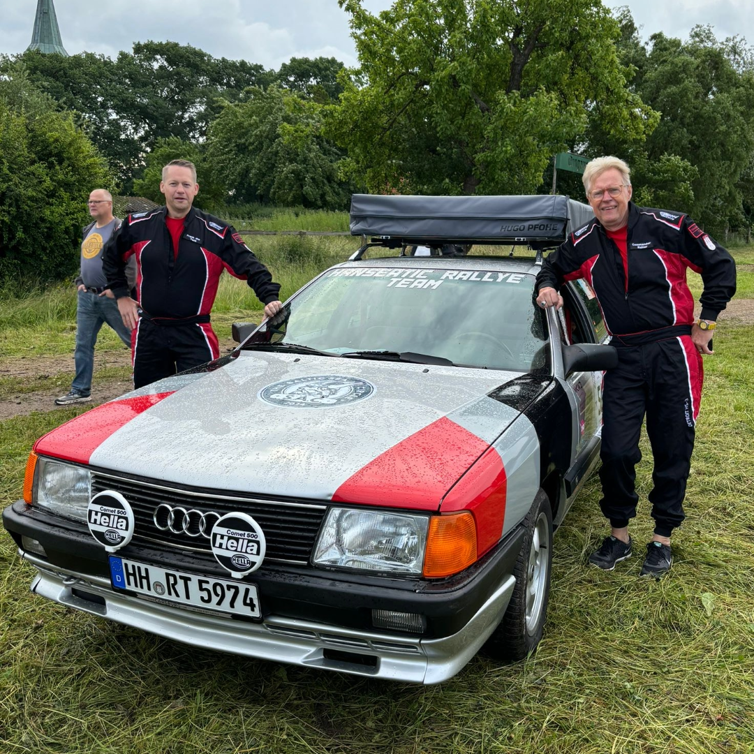 Zwei Männer in Rallyeanzügen stehen neben einem Audi mit Rallye-Design auf einer Wiese. Das Auto hat Aufkleber und Zusatzscheinwerfer. Ein weiteres Auto befindet sich im Hintergrund.