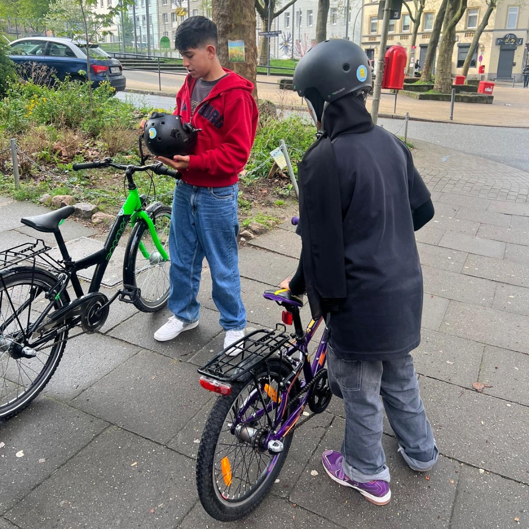 Zwei Kinder mit Fahrrädern halten Fahrradhelme, eines trägt bereits einen Helm.
