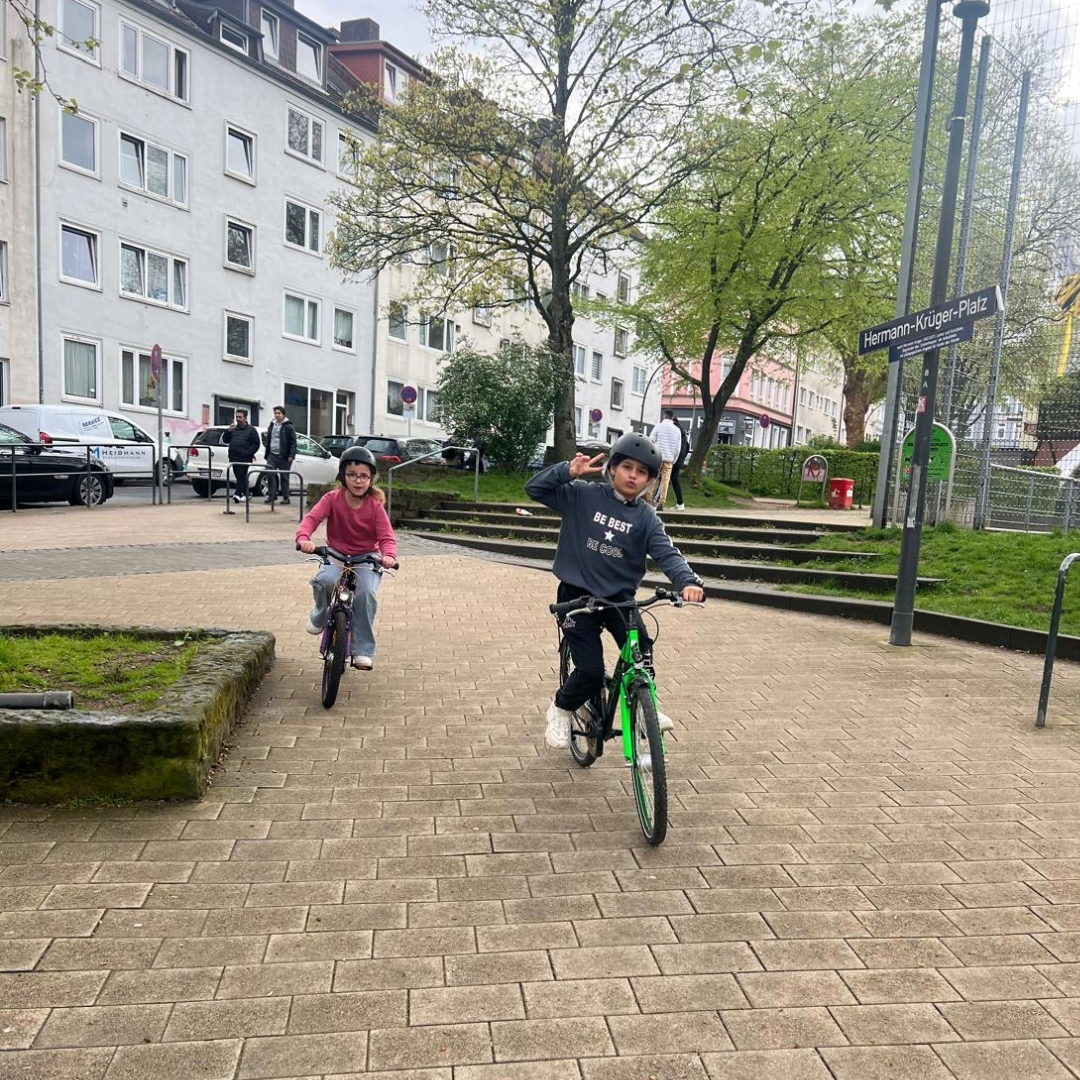 Zwei Kinder fahren auf einem Platz Fahrrad, eines zeigt ein Peace-Zeichen, Schilder markieren den Hermann-Krüger-Platz.