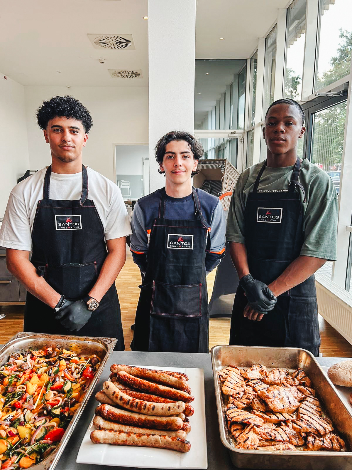 3 Jugendliche stehen in Schürzen gekleidet hinter diversem Grillgut.