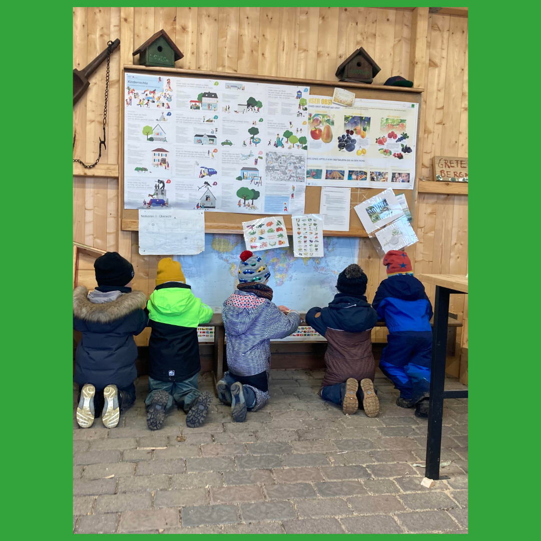 Kinder sitzen auf dem Boden und betrachten eine mit Lernmaterialien und Karten gefüllte Pinnwand.