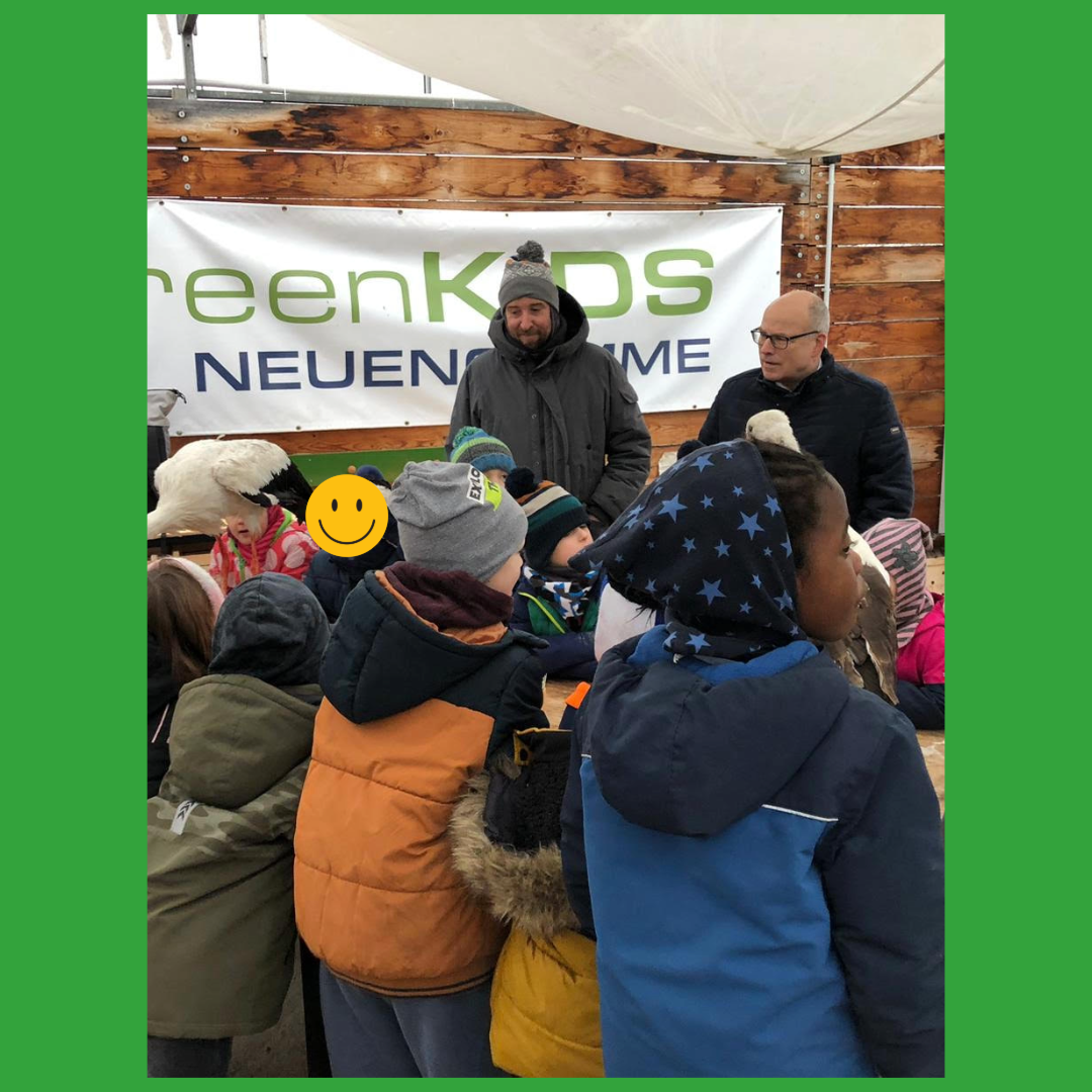Kinder und Erwachsene stehen vor einem Banner mit der Aufschrift "GreenKIDS Neuengamme" und unterhalten sich.