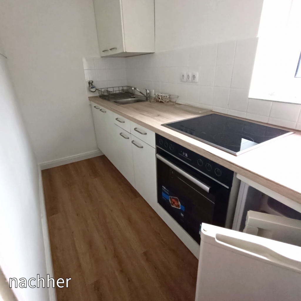 Moderner, schmaler Küchenbereich mit Holzfußboden, weißen Schränken und einem eingebauten Elektroherd. Spüle und Backofen integriert; ein Fenster sorgt für Licht. Text 