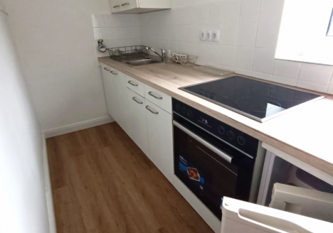 Moderner, schmaler Küchenbereich mit Holzfußboden, weißen Schränken und einem eingebauten Elektroherd. Spüle und Backofen integriert; ein Fenster sorgt für Licht. Text "nachher" in der Ecke.