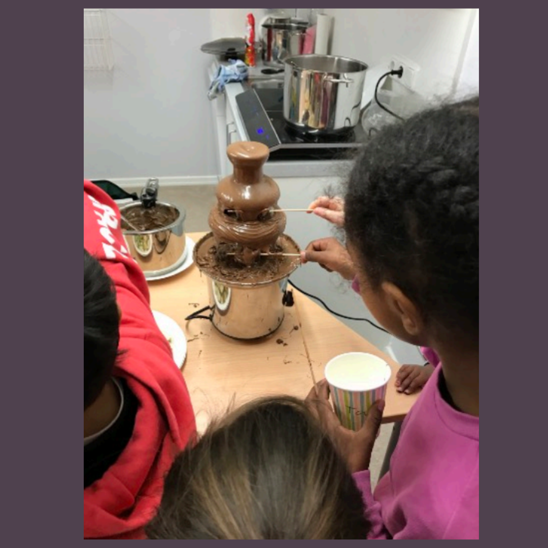 Kinder stehen um einen Schokoladenbrunnen. Sie tauchen Spieße in die fließende Schokolade. Im Hintergrund befindet sich eine Küche mit Töpfen und einem Herd.