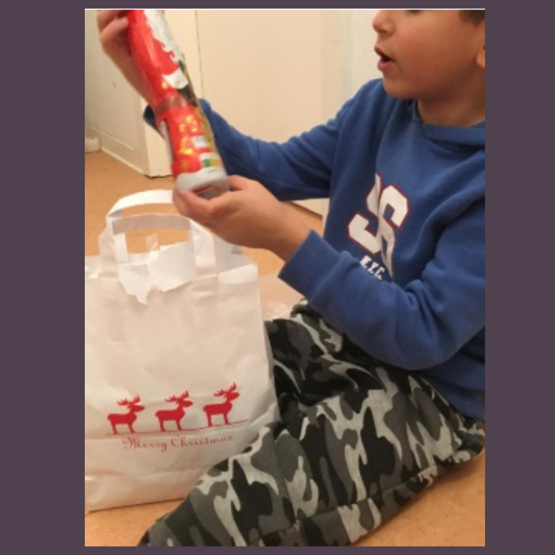 Ein Kind in einem blauen Hoodie und Tarnmusterhose sitzt auf dem Boden und hält einen Schokoladen-Weihnachtsmann. Neben ihm steht eine weiße Tüte mit roten Rentieren und "Merry Christmas"-Aufdruck.