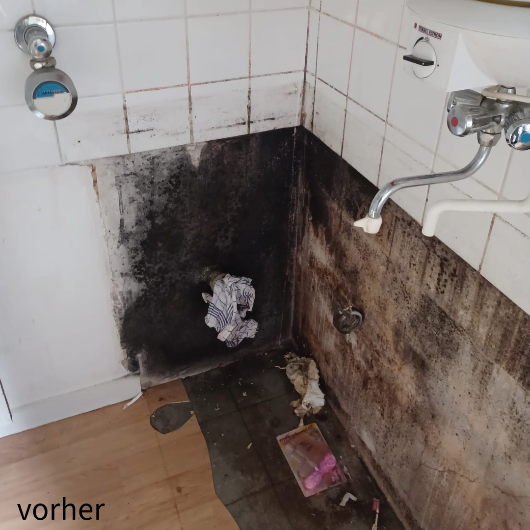 Ein stark verschmutzter Wand- und Bodenbereich einer Küche mit dunklen Schimmelspuren und einem unbenutzten Wasseranschluss. Der Boden ist teilweise dreckig. Text "vorher" in der Ecke.